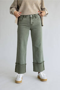 A person wearing olive green wide-leg cropped jeans with rolled cuffs and raw fringe, styled with a beige sweater and white platform sneakers against a light gray studio background.