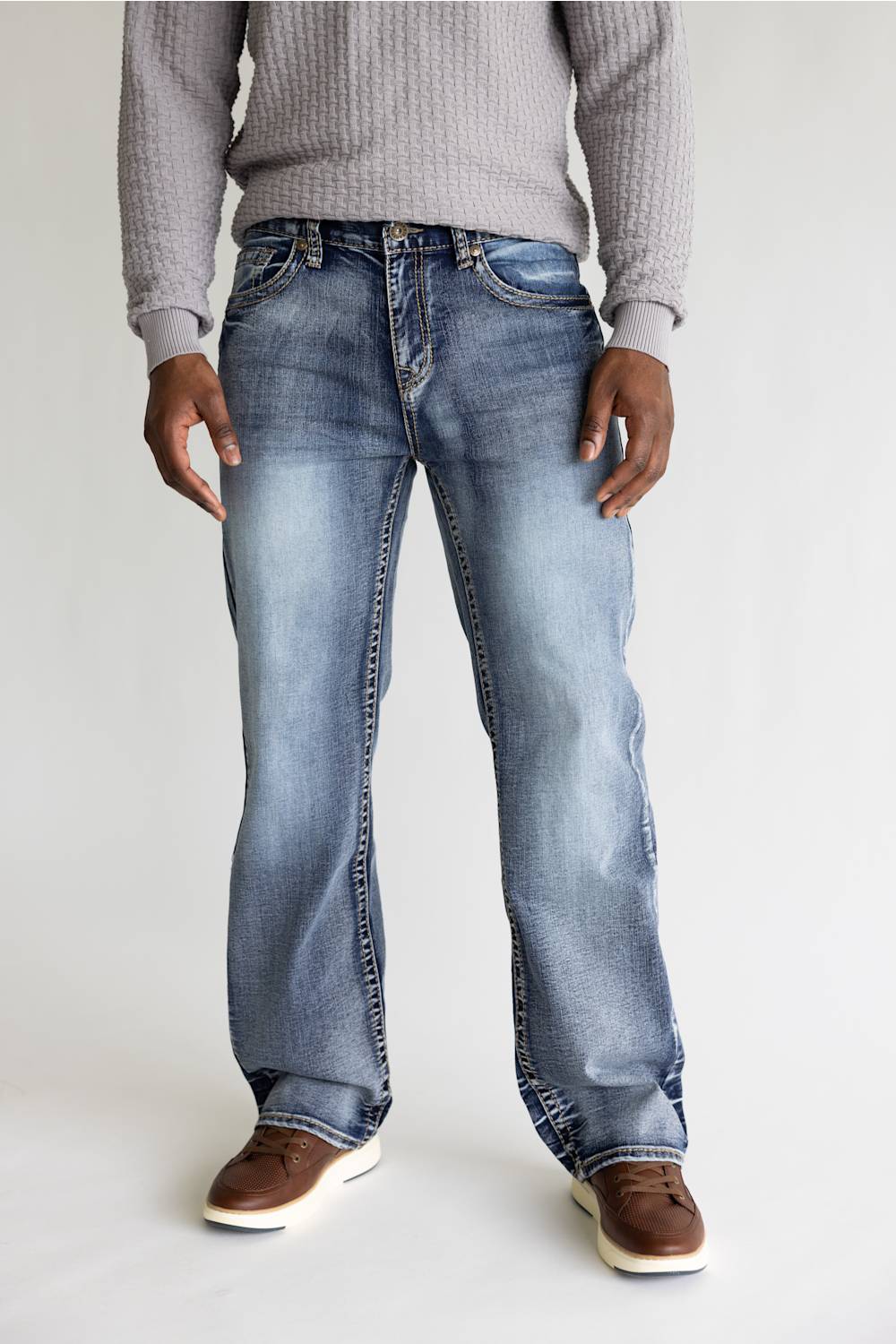 A person wearing light-wash blue jeans with a gray ribbed sweater and brown shoes, standing against a plain light gray studio background.