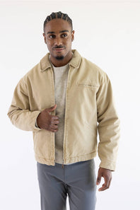 A man wearing a beige canvas utility jacket over a light gray sweater and gray pants, standing against a plain white studio background.