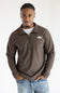 A man wearing a brown quarter-zip fleece pullover by The North Face, styled over a white t-shirt and blue jeans, standing against a plain white background.