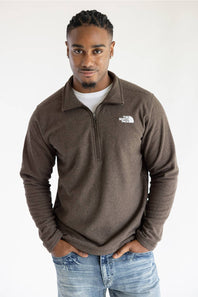 A man wearing a brown quarter-zip fleece pullover by The North Face, styled over a white t-shirt and blue jeans, standing against a plain white background.