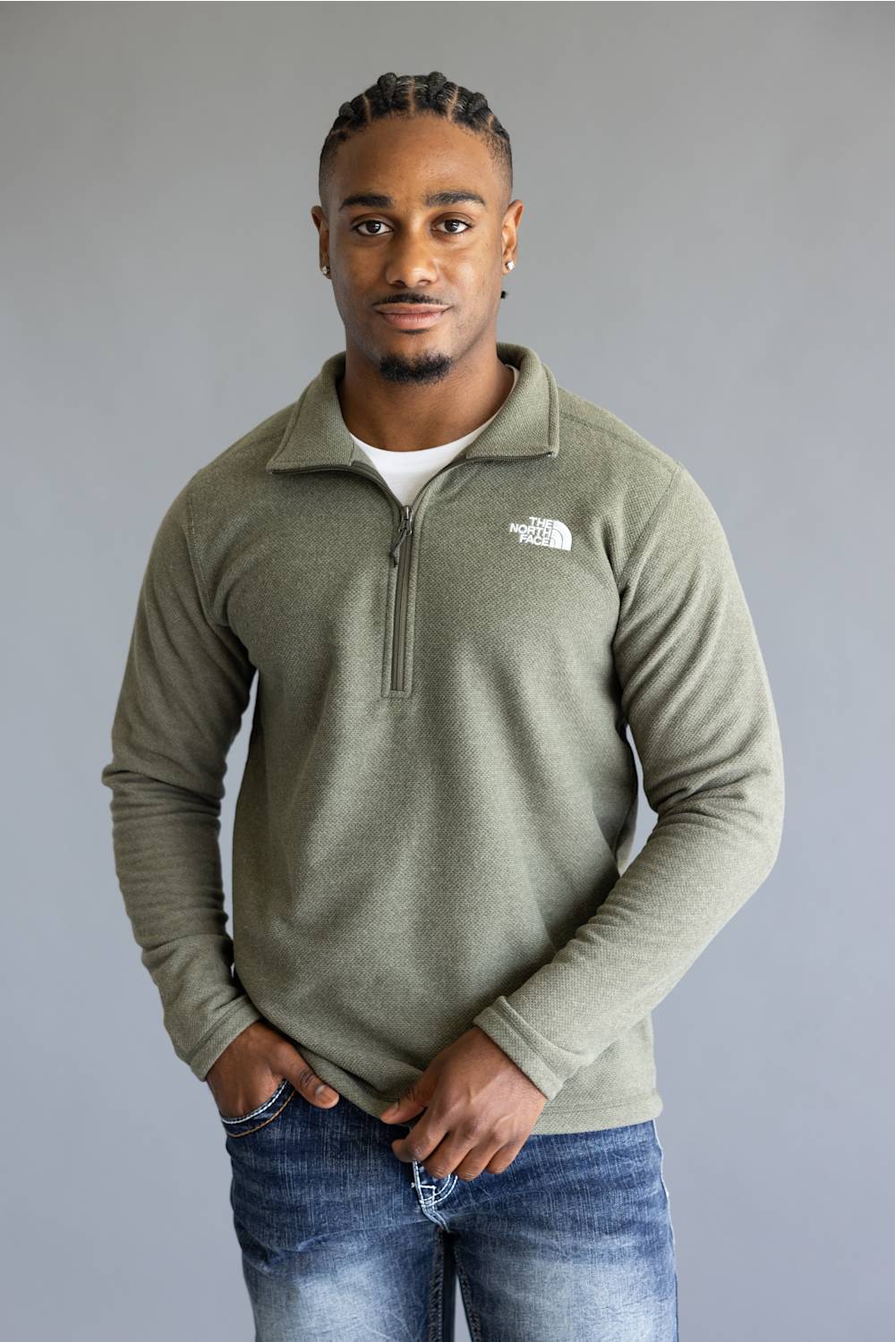 A man wearing an olive green quarter-zip fleece pullover by The North Face, paired with blue jeans, standing against a solid gray studio background.