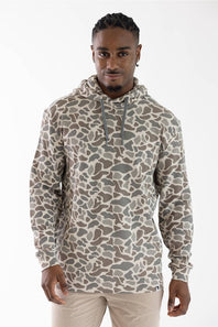 A man wearing a camouflage-patterned pullover hoodie in shades of beige, brown, and gray, paired with beige pants, photographed against a plain white studio background.