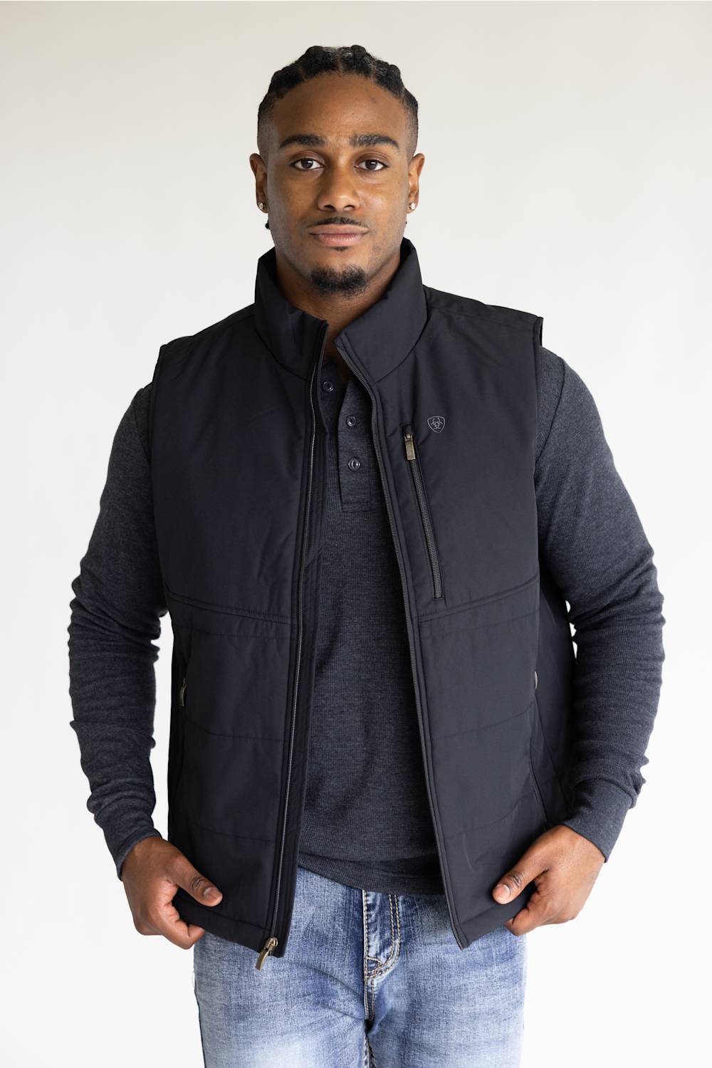 A man wearing a black zip-up insulated vest over a dark gray long-sleeve shirt and blue jeans, standing against a plain light background.