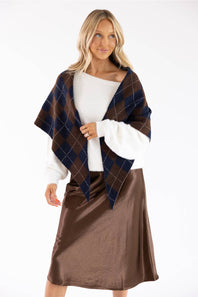 A woman modeling a brown and navy blue argyle plaid wrap or poncho draped over a white sweater and brown satin skirt, against a plain white studio background.