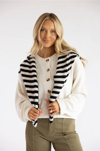 A blonde woman modeling a cream knit cardigan with black and white stripes on the shoulders, worn over a cream button-up sweater and olive green pants, against a plain white studio background.