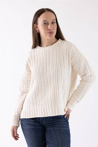 A woman wearing a cream-colored ribbed knit sweater and dark blue jeans, standing against a plain white studio background.