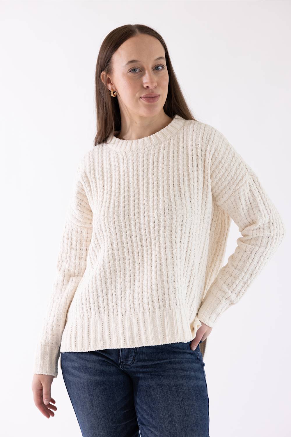 A woman wearing a cream-colored ribbed knit sweater and dark blue jeans, standing against a plain white studio background.