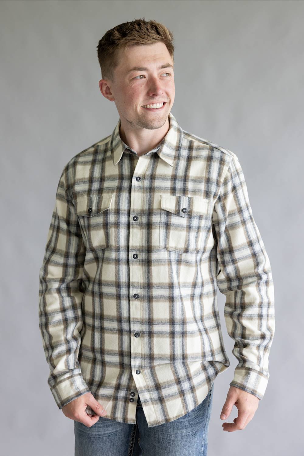 Front view of a smiling male model with hands in his pockets wearing the Split Flannel Shirt for Men in Grey Ecru, featuring a beige, gray, and brown plaid pattern, long sleeves, and dual flap chest button pockets, paired with medium-wash jeans against a neutral gray studio background.