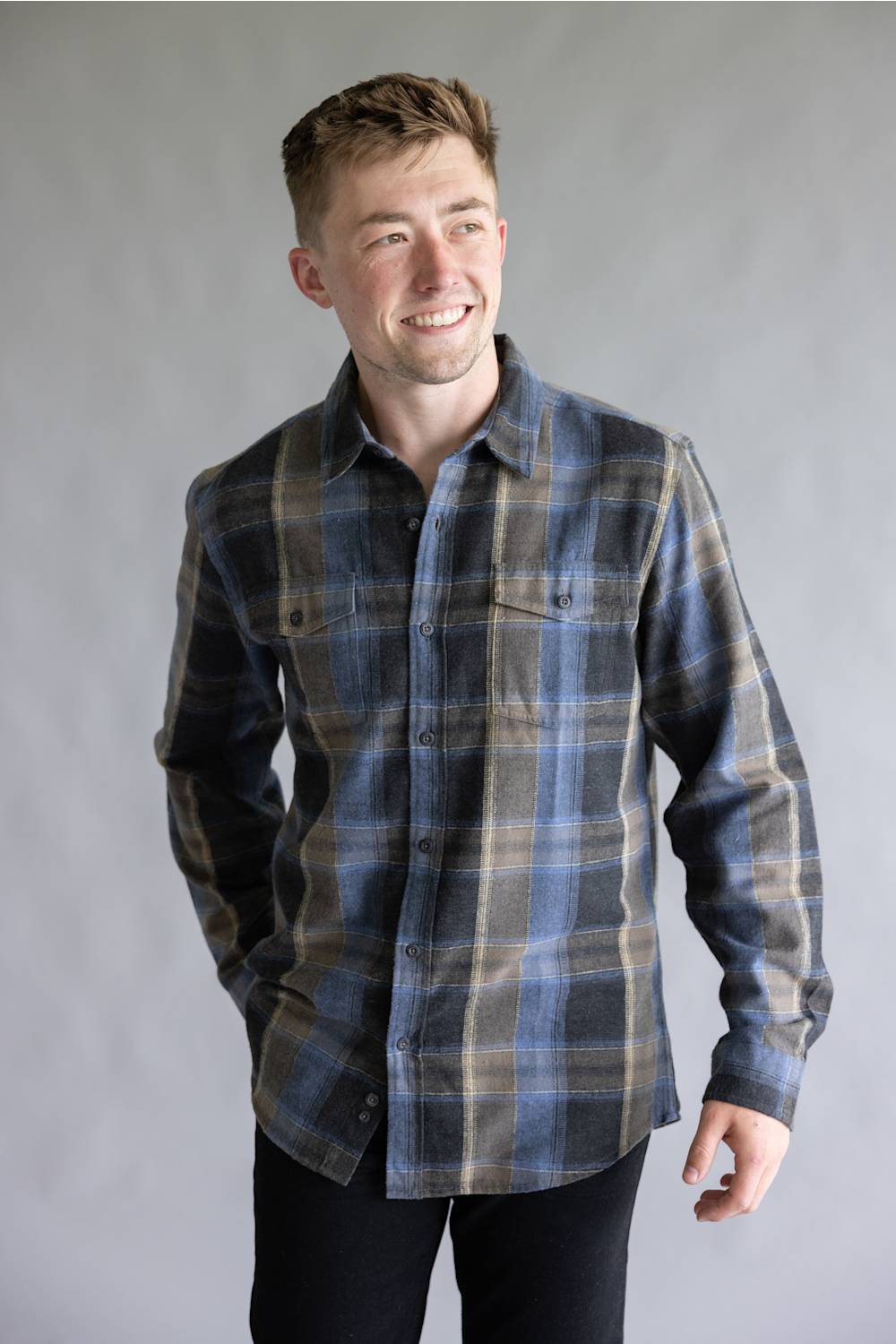 A smiling male model posing against a neutral gray studio background, wearing the Split Flannel Shirt for Men in Moka featuring a blue, dark gray, and tan plaid pattern with long sleeves and dual buttoned chest pockets.
