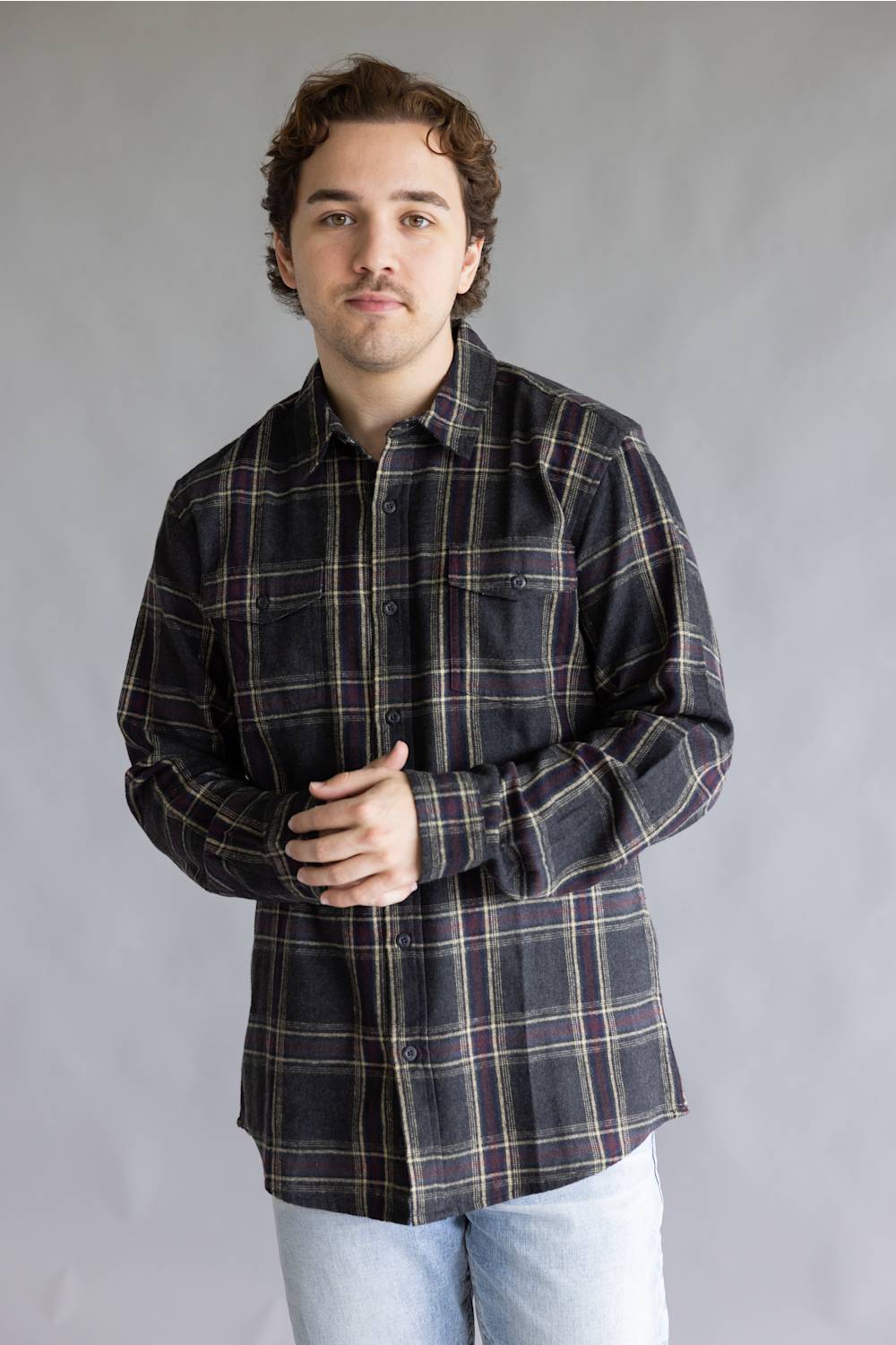 Front view of a male model with hands clasped in front of him wearing the Split Flannel Shirt for Men in Slate, showcasing the charcoal gray plaid pattern with burgundy and cream accent lines, long sleeves, dual buttoned chest pockets, and light wash jeans against a plain gray studio background.