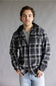 Model wearing the Split Men's long-sleeve hooded flannel shirt in a black, grey, and white plaid pattern, featuring two chest flap pockets and a solid black attached hood against a grey studio background.