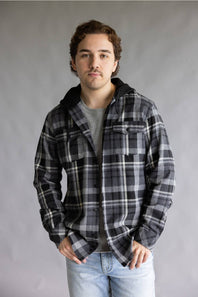 Model wearing the Split Men's long-sleeve hooded flannel shirt in a black, grey, and white plaid pattern, featuring two chest flap pockets and a solid black attached hood against a grey studio background.