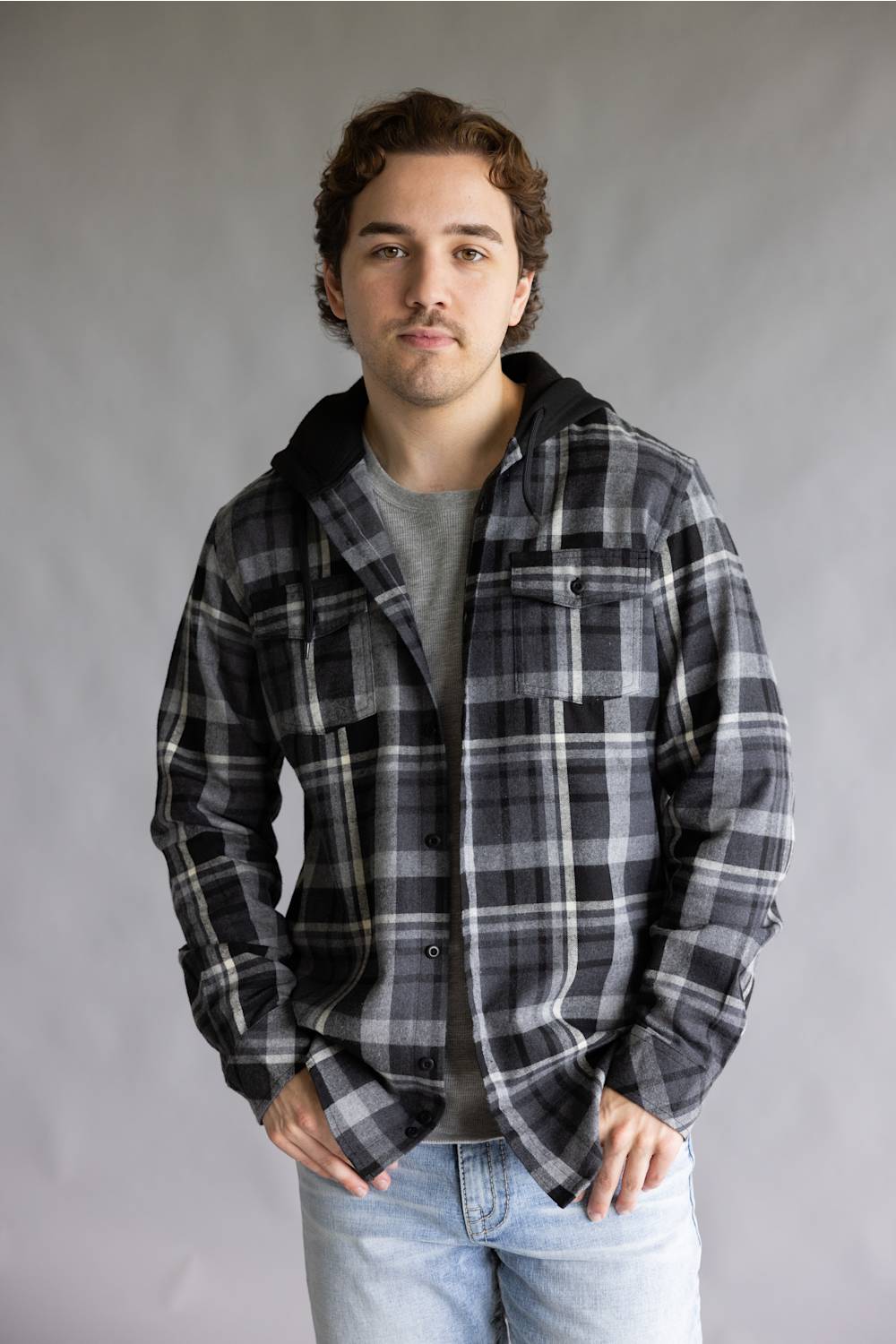 Model wearing the Split Men's long-sleeve hooded flannel shirt in a black, grey, and white plaid pattern, featuring two chest flap pockets and a solid black attached hood against a grey studio background.