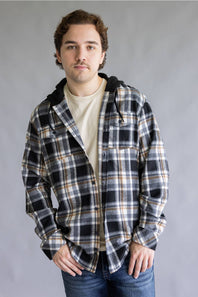 Model wearing an open, unbuttoned Split Men's long-sleeve hooded flannel shirt in a cream, black, and tan plaid pattern, styled over a plain cream t-shirt and blue jeans with hands in pockets against a gray studio background.