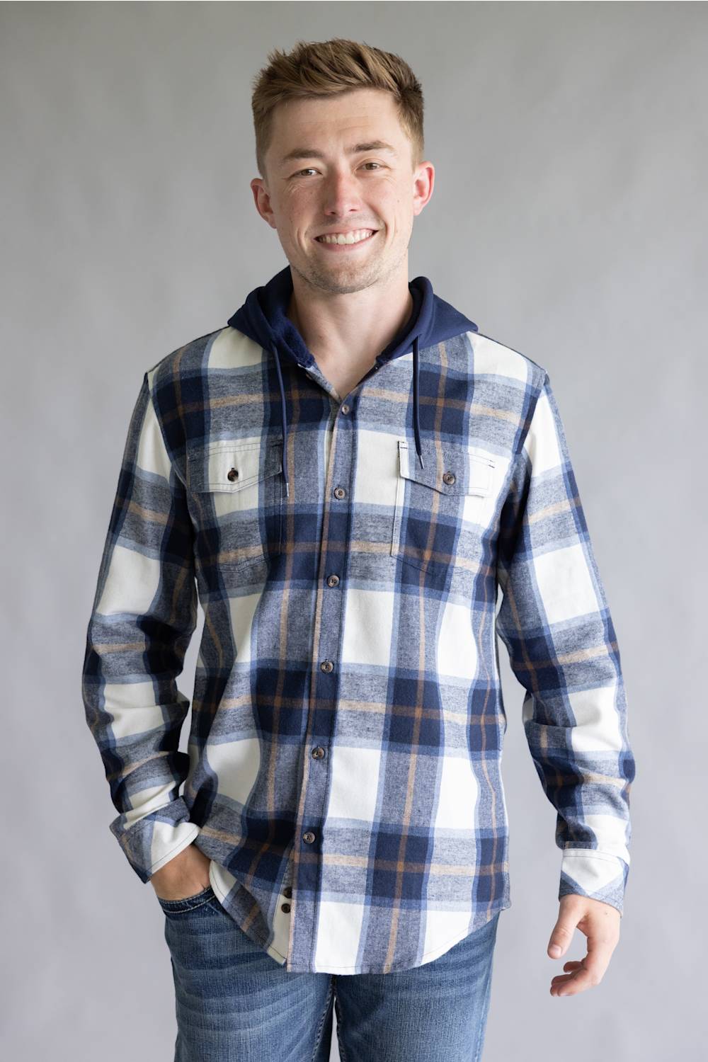 Model wearing the Split Men's long-sleeve hooded flannel shirt in a navy, ecru white, and tan plaid pattern, featuring two chest flap pockets and a solid navy attached hood with drawstrings against a gray studio background.