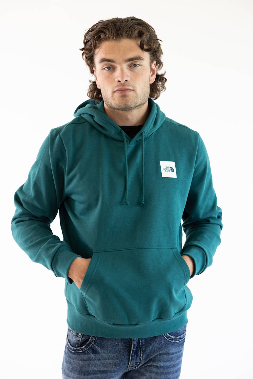A man wearing a teal green pullover hoodie with a white square The North Face logo patch on the left chest, paired with blue jeans, photographed against a plain white studio background.
