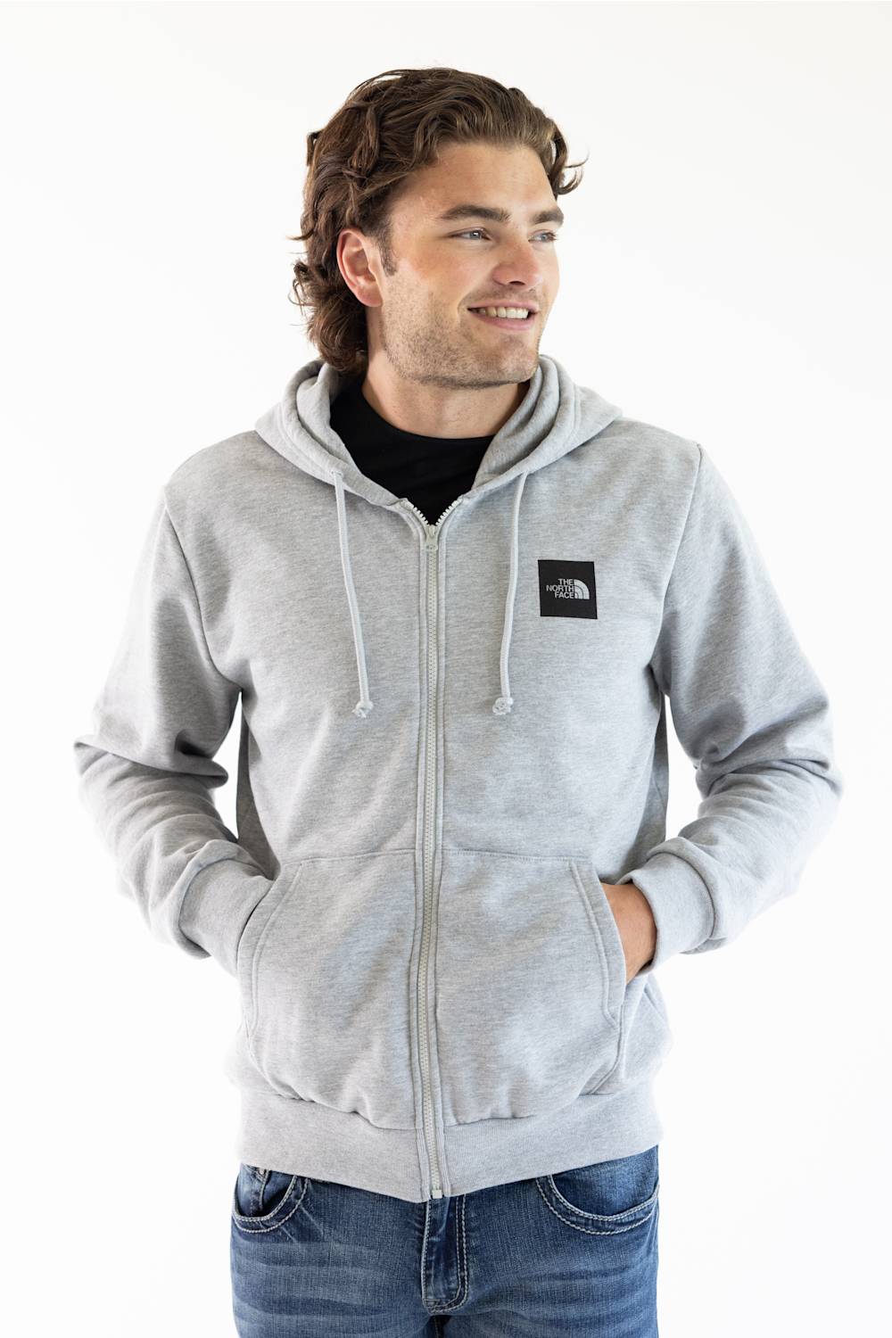 A man wearing a light gray zip-up hoodie with a black chest logo patch, paired with a black t-shirt and blue jeans, photographed against a plain white background.