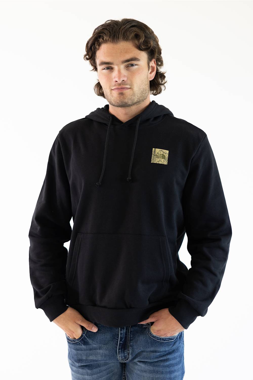A man wearing a black pullover hoodie with a small square graphic patch on the left chest, paired with blue jeans, photographed against a plain white background.