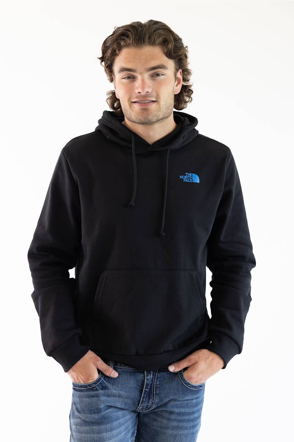 A man wearing a black pullover hoodie with a small blue The North Face logo on the left chest, paired with blue jeans, against a plain white background.