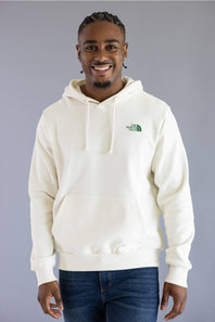 A man wearing a cream-colored pullover hoodie with a green embroidered logo on the left chest, paired with dark blue jeans, standing against a solid gray studio background.