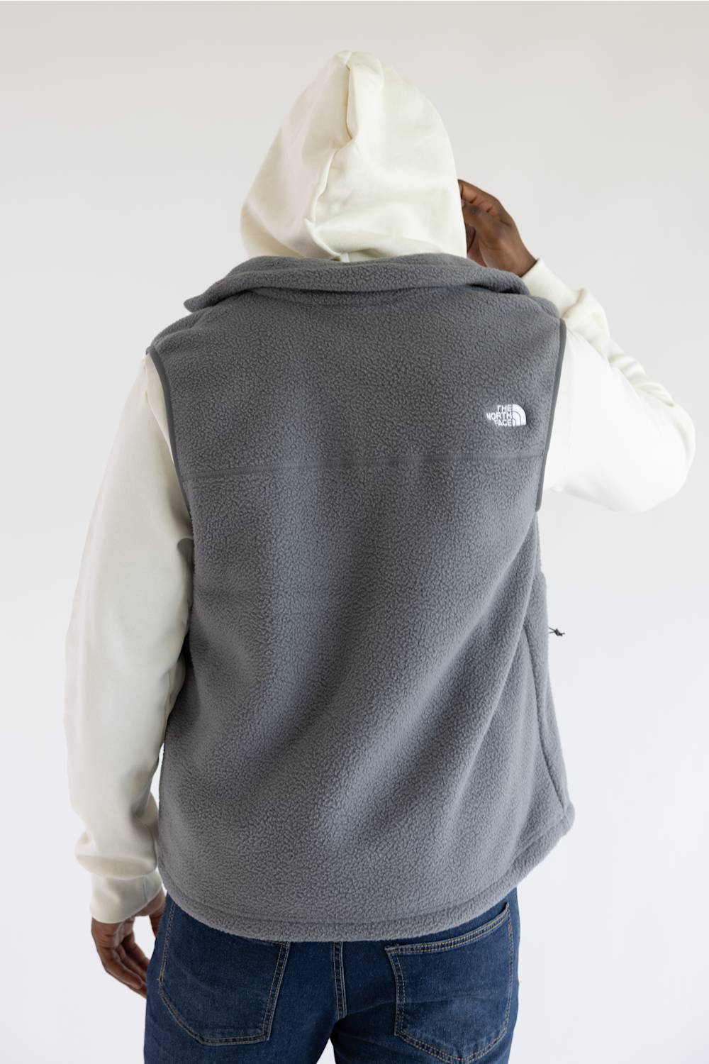 A person shown from the back wearing a gray fleece vest over a cream hoodie and blue jeans, with The North Face logo visible on the upper back.