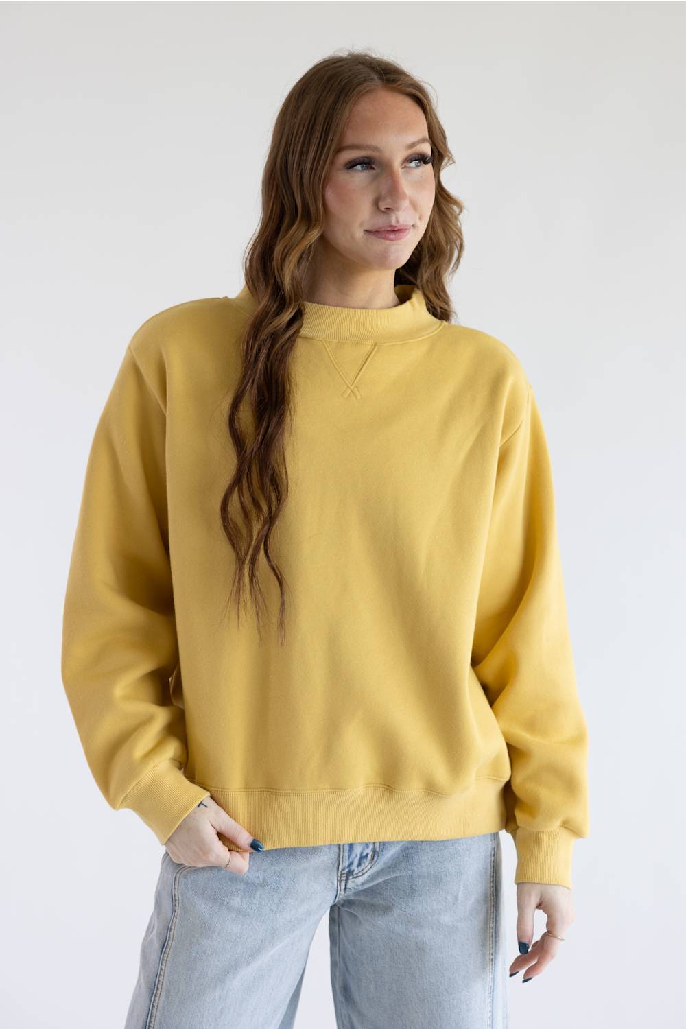 A woman with long brown hair wearing a mustard yellow crewneck sweatshirt and light blue jeans, standing against a plain white studio background.