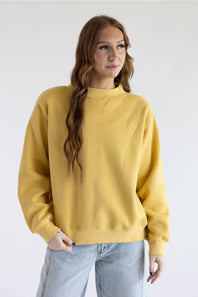 A woman with long brown hair wearing a mustard yellow crewneck sweatshirt and light blue jeans, standing against a plain white studio background.