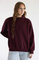 A woman with red hair wearing a loose-fitting, crewneck knit sweater in deep burgundy and blue jeans, standing against a plain white studio background.