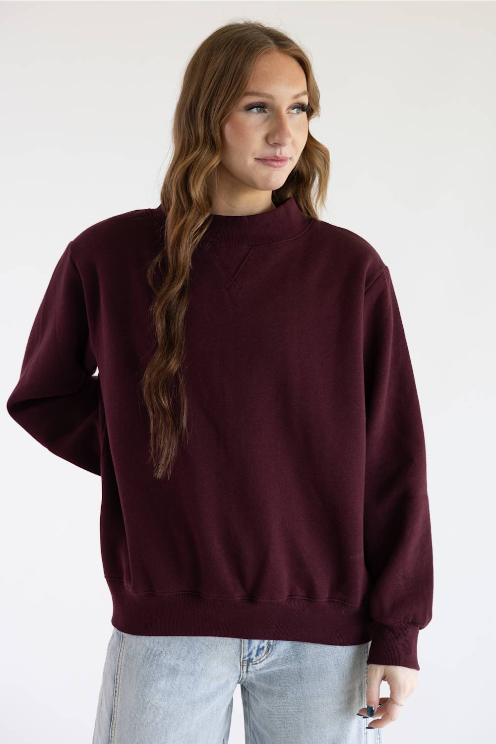 A woman with red hair wearing a loose-fitting, crewneck knit sweater in deep burgundy and blue jeans, standing against a plain white studio background.