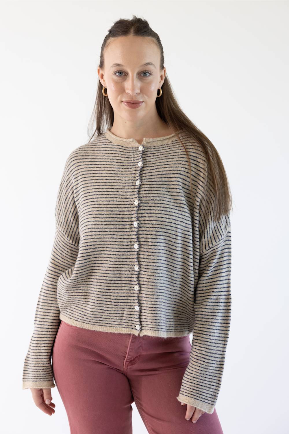 A woman modeling a cropped, long-sleeve knit cardigan with horizontal stripes in beige, featuring a button-down front. She is wearing dusty rose pants against a plain white studio background.