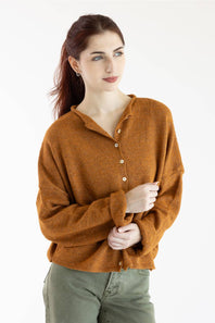 A woman wearing a warm rust-orange knit button-front cardigan sweater with a split neckline, paired with olive green pants, photographed against a clean white studio background.