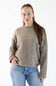 A woman modeling a light brown knit crewneck sweater with blue jeans against a plain white background.