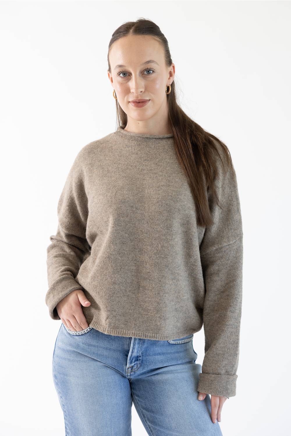 A woman modeling a light brown knit crewneck sweater with blue jeans against a plain white background.