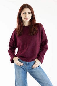 A woman with red hair wearing a loose-fitting, crewneck knit sweater in deep burgundy and blue jeans, standing against a plain white studio background.