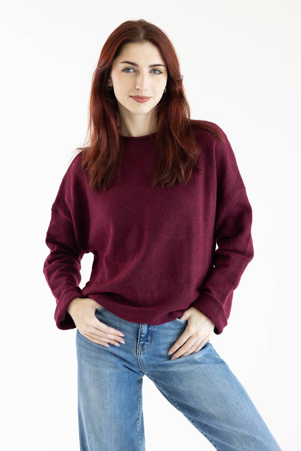 A woman with red hair wearing a loose-fitting, crewneck knit sweater in deep burgundy and blue jeans, standing against a plain white studio background.