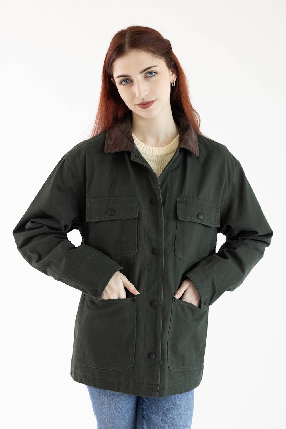 A woman with red hair wearing an oversized dark olive green utility jacket with a brown collar, over a cream sweater and blue jeans, photographed against a plain white background.