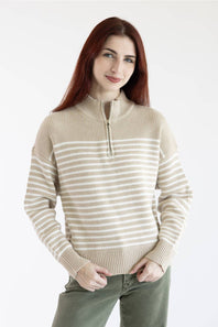 A woman with long red hair wearing a beige and white horizontally striped quarter-zip sweater and olive green pants, standing against a plain white studio background.