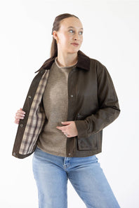 A woman modeling a dark brown leather jacket with a brown plaid lining, worn open over a beige sweater and blue jeans, against a plain white background.