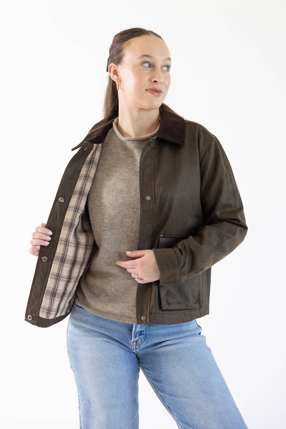 A woman modeling a dark brown leather jacket with a brown plaid lining, worn open over a beige sweater and blue jeans, against a plain white background.