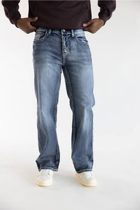 A person wearing light-to-medium wash blue jeans with a straight-leg fit, paired with a dark brown long-sleeve top and white sneakers, shown from the waist down on a light gray studio background.