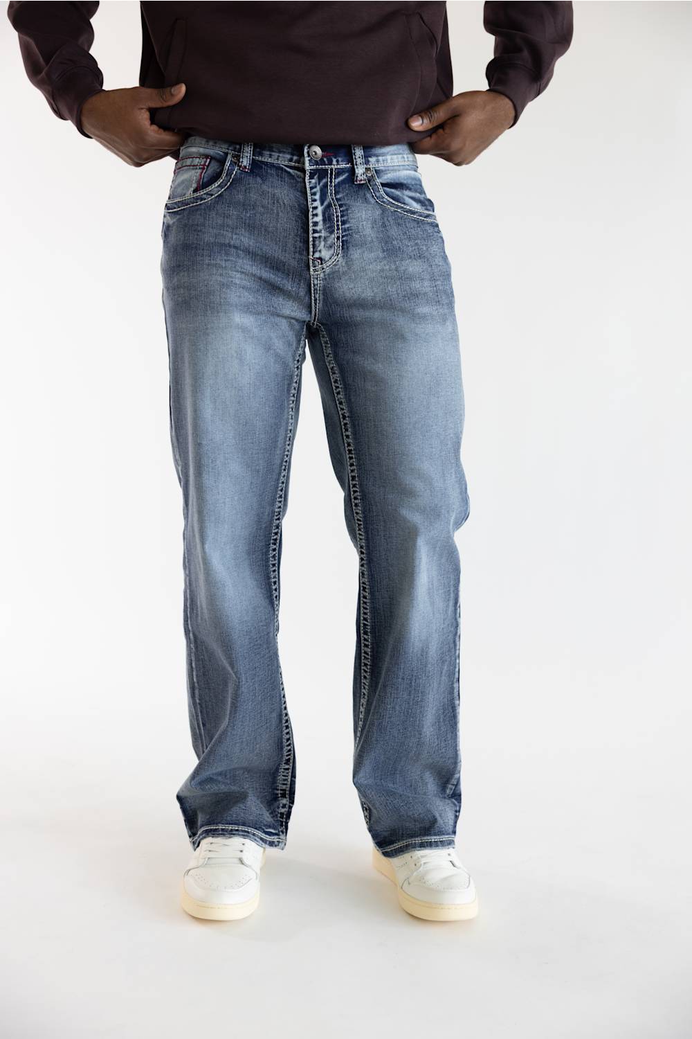 A person wearing light-to-medium wash blue jeans with a straight-leg fit, paired with a dark brown long-sleeve top and white sneakers, shown from the waist down on a light gray studio background.
