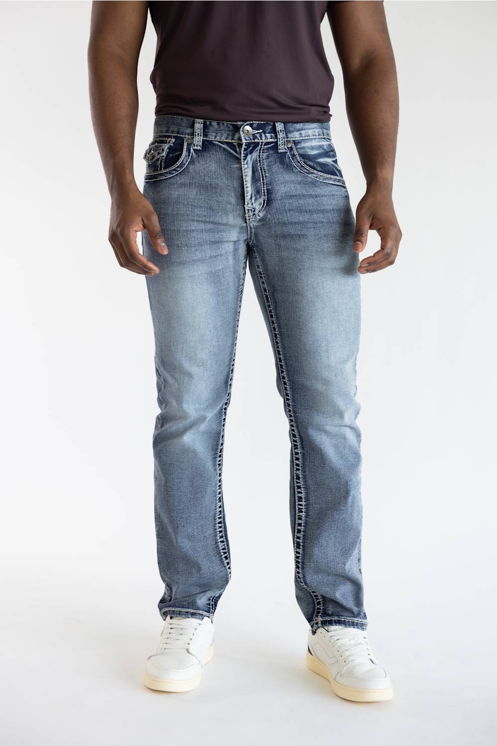 A person wearing light-wash blue denim jeans with white sneakers and a dark top, standing against a plain light gray studio background.