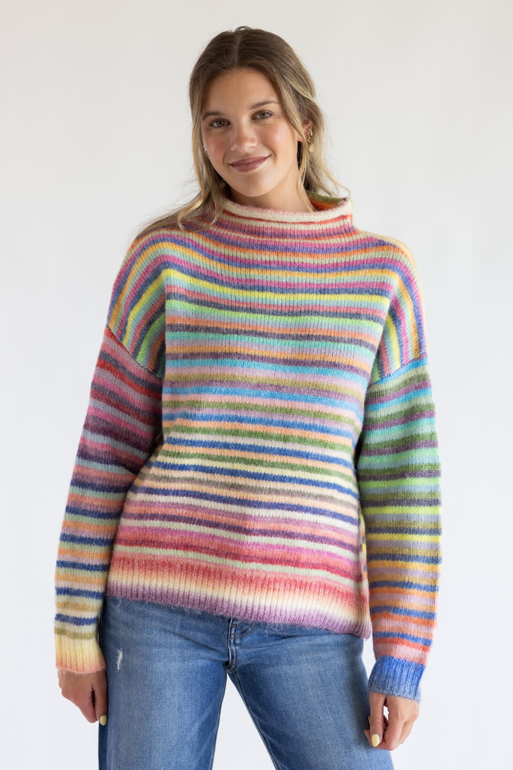 Woman wearing a striped multi rainbow colored mock neck sweater with blue denim jeans standing in front of a clean white background