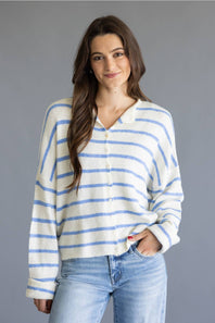 A woman wearing a cream-colored knit cardigan with thin blue horizontal stripes, paired with light-wash blue jeans, against a solid gray studio background.