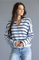 A woman wearing a white knit button-up cardigan with navy blue horizontal stripes, paired with light-wash blue jeans, against a solid gray studio background.