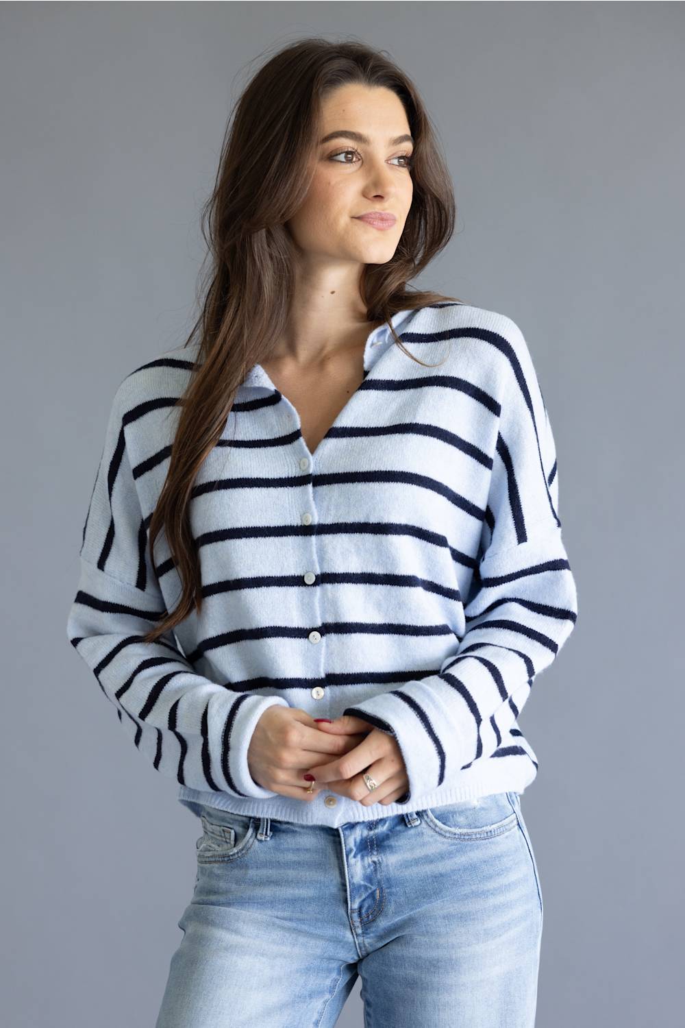 A woman wearing a white knit button-up cardigan with navy blue horizontal stripes, paired with light-wash blue jeans, against a solid gray studio background.
