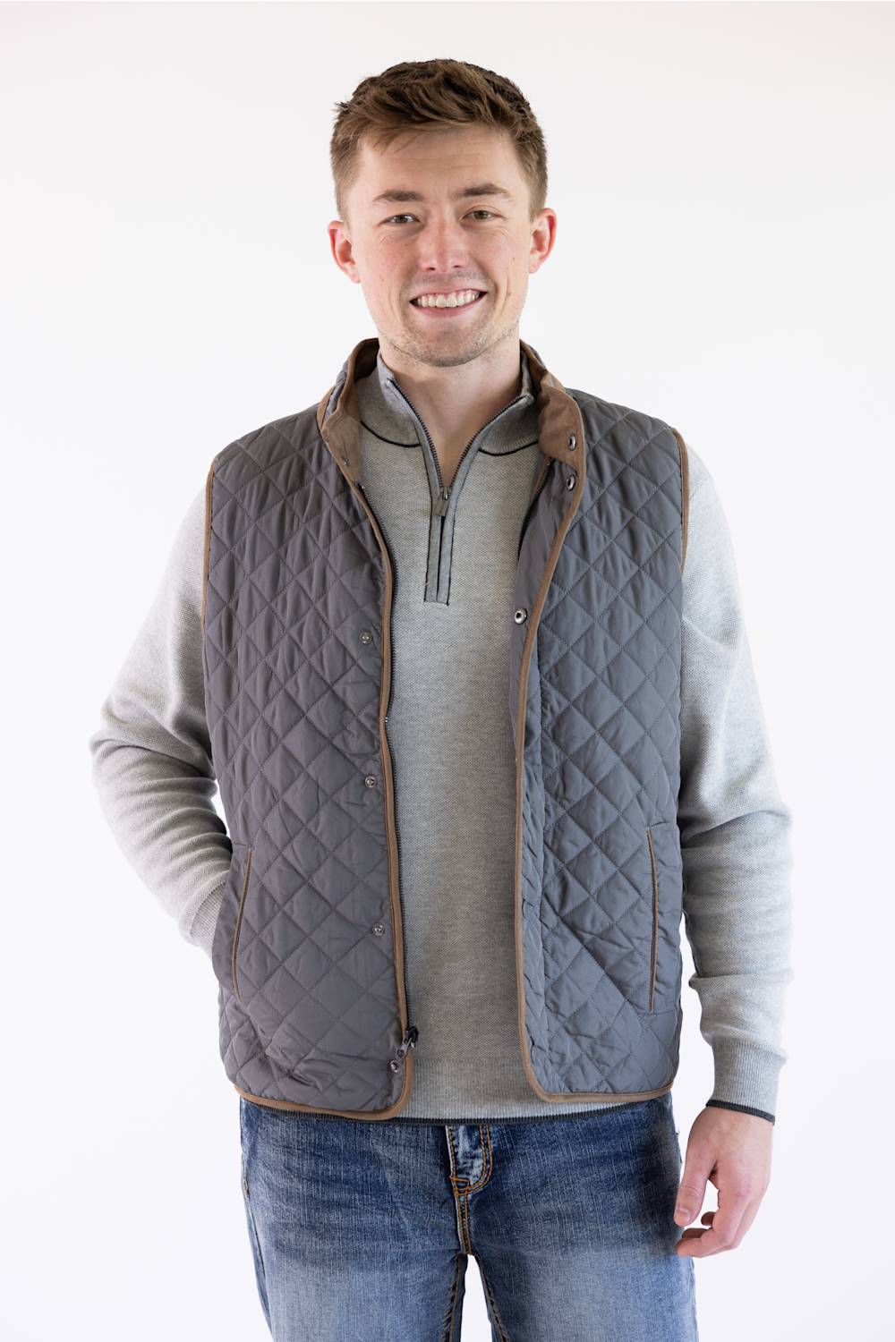 A male model wearing the FX Fusion Upland Quilted Field Vest in charcoal over a grey mock-neck quarter-zip sweater and blue jeans, standing against a neutral light background with his right hand in his pocket.
