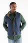 A man wearing a navy blue quilted vest with tan trim over a green button-up shirt and blue jeans, photographed against a plain white studio background.