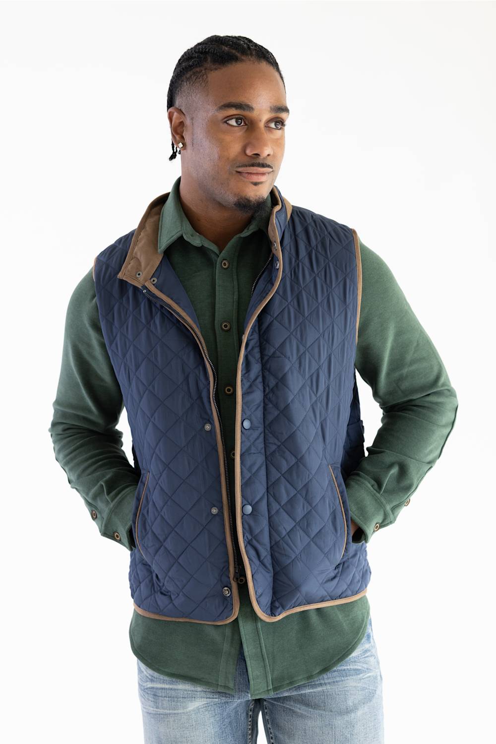 A man wearing a navy blue quilted vest with tan trim over a green button-up shirt and blue jeans, photographed against a plain white studio background.